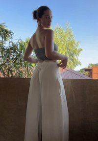 Femme portant un top tube vert et un pantalon blanc à jambes larges, debout sur un balcon, regardant par-dessus son épaule avec de la verdure et des toits en arrière-plan.