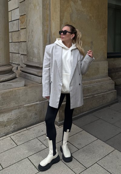 Blazer gris claro de corte cruzado, sudadera blanca, leggins negros y botas blancas de suela gruesa con acentos negros; todo ello frente a una pared de piedra texturada.