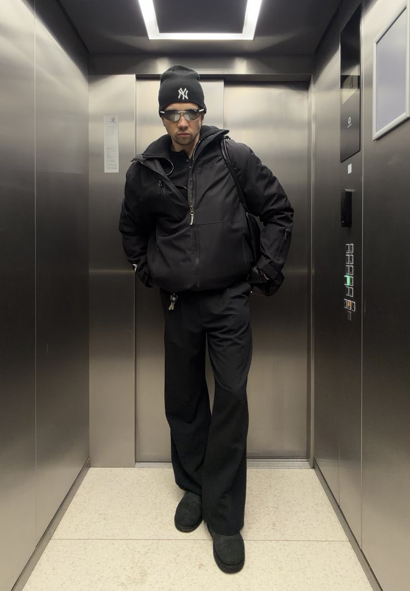 Homme portant une veste noire, pantalon large, bonnet, lunettes de soleil et gants, debout dans un ascenseur en métal avec des boutons éclairés sur le panneau droit.