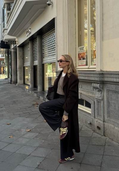 Mujer con un abrigo largo y oscuro ajusta su zapato mientras está de pie en una acera de la ciudad frente a una tienda COS cerrada con fachada de piedra gris.