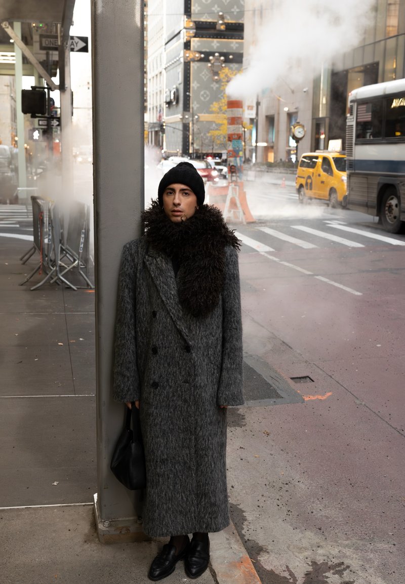 Personne en manteau gris long, bonnet noir et grande écharpe marron, debout contre un poteau de rue en ville avec de la vapeur et de la circulation en arrière-plan.