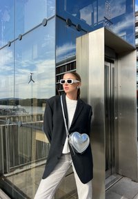 Kvinna i en överdimensionerad svart blazer och vita solglasögon står vid en glaselevator med en metallisk hjärtformad handväska över kroppen.