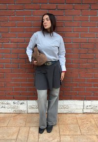 Γυναίκα με καστανά μαλλιά στέκεται μπροστά σε κόκκινο τοίχο από τούβλα, φοράει γκρι πουλόβερ πάνω από λευκό πουκάμισο, σκούρα φούστα πάνω από γκρι τζιν, κρατώντας καφέ τσάντα.