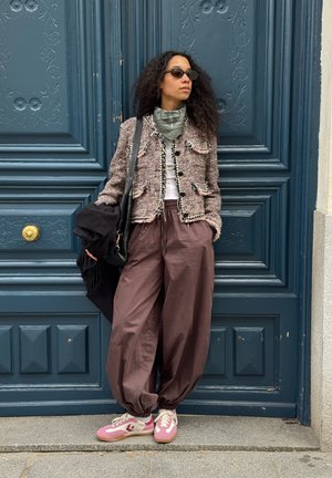 Mujer con cabello rizado que lleva gafas de sol, chaqueta de tweed, bandana verde, pantalones marrones sueltos y zapatillas rosas, de pie frente a una puerta azul tallada con ornamentos.