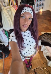 Jeune femme aux longs cheveux violets portant un haut blanc à pois et un bandeau assorti, debout dans une pièce où des vêtements et des chaussures sont visibles.