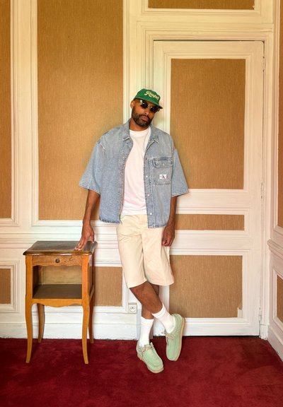 Camisa de denim oversized, camiseta blanca, pantalones cortos beige, zapatos verde menta y gorra verde. Mesita de noche de madera con un cajón, fondo de alfombra roja.