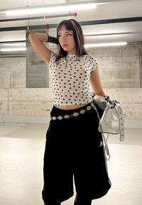 Top cu mânecă scurtă, cu buline alb-negru; pantaloni scurți negri, largi; curea argintie; geantă argintie strălucitoare; fundal de beton cu lumini deasupra.