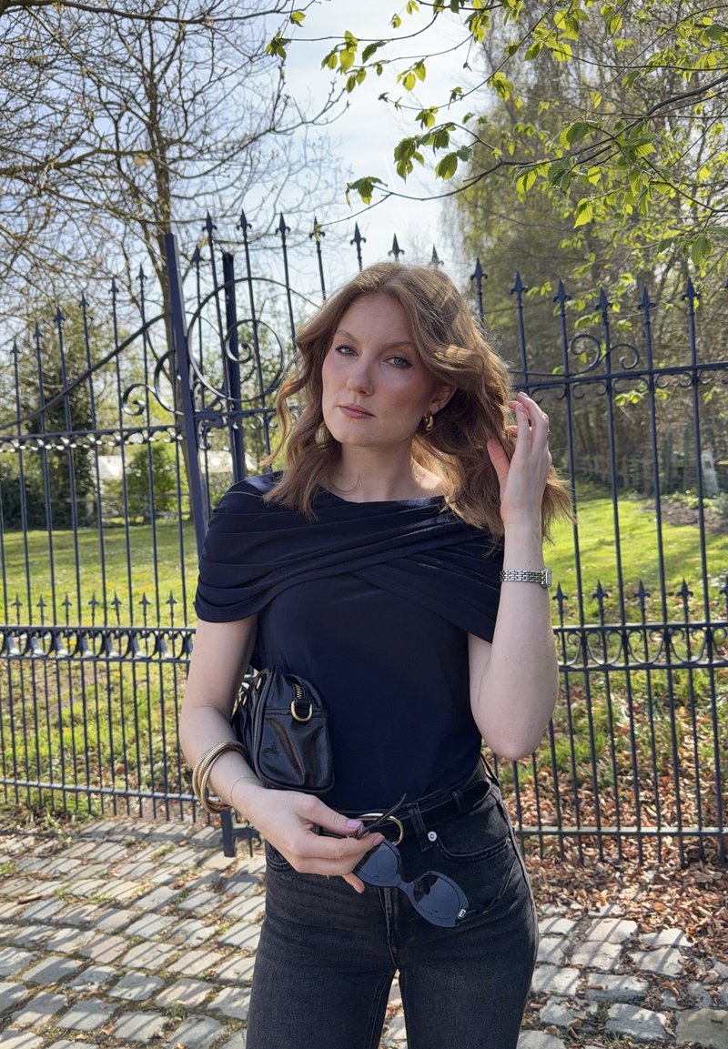 Jonge vrouw met golvend haar, gekleed in een zwart topje en jeans, houdt een zonnebril en een kleine tas vast, staand bij een smeedijzeren hek in een park.