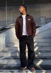 Άνδρας στέκεται σε σκάλες από σκυρόδεμα με τα χέρια στις τσέπες, φορώντας καφέ μπουφάν, λευκό πουκάμισο, μαύρο παντελόνι και καφέ παπούτσια, κοιτάζοντας προς τα αριστερά.