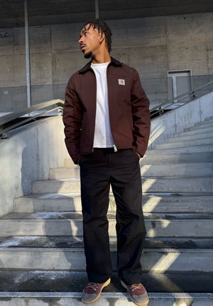 Hombre de pie en escaleras de concreto con las manos en los bolsillos, vistiendo chaqueta marrón, camisa blanca, pantalones negros y zapatos marrones, mirando hacia la izquierda.