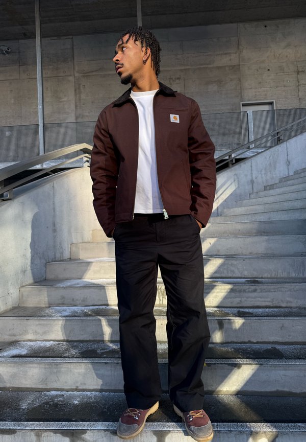 Man stands on concrete steps with hands in pockets, wearing a brown jacket, white shirt, black trousers, and brown shoes, looking to the left.