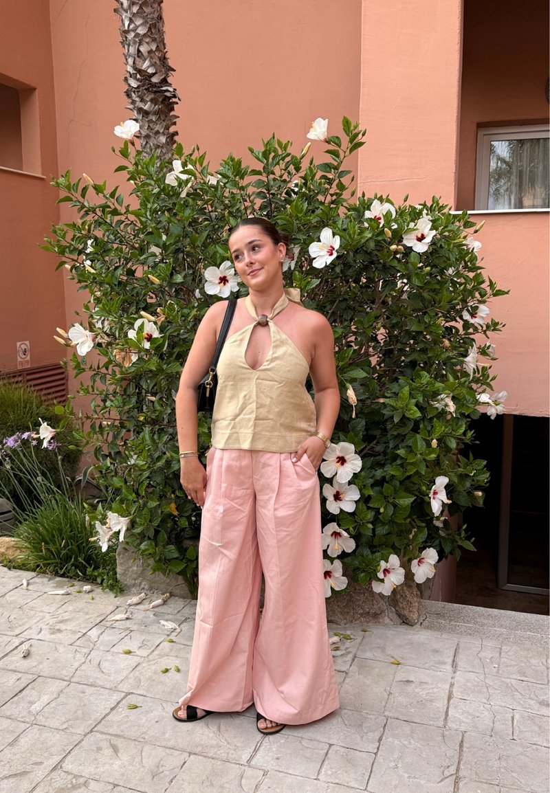 Jeune femme en haut beige à col halter et pantalon large rose debout près d’un buisson d’hibiscus en fleurs près d’un bâtiment couleur pêche.