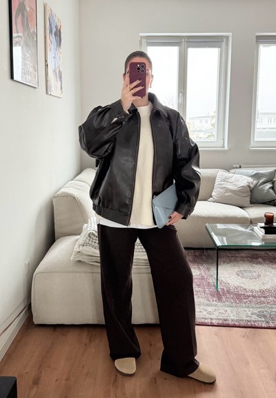 Chaqueta de cuero negra con cremallera y mangas abullonadas, suéter blanco, pantalones de pierna ancha marrones y cartera azul clara en un acogedor entorno interior.