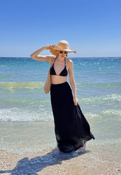 Top de bikini negro combinado con una falda larga negra y fluida, sombrero de paja y bolso de hombro tejido, de pie junto a la orilla del agua.
