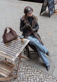 Jaqueta de couro castanha com colarinho de estampa de leopardo, luvas pretas, suéter escuro, jeans azuis flare, botas pretas de bico fino, bolsa castanha e uma chávena de café sobre uma mesa de madeira.