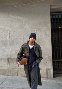 Άνδρας που φοράει ένα σκούρο σκουφάκι και ένα μακρύ πράσινο παλτό, κρατώντας μια καφέ δερμάτινη τσάντα, που στέκεται δίπλα σε έναν πέτρινο τοίχο με μια μαύρη μεταλλική πύλη κοντά.