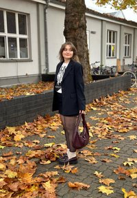 Donna che indossa un blazer nero, una gonna, collant, con una borsa bordeaux in mano, in piedi su un marciapiede coperto di foglie autunnali vicino a un albero e biciclette.