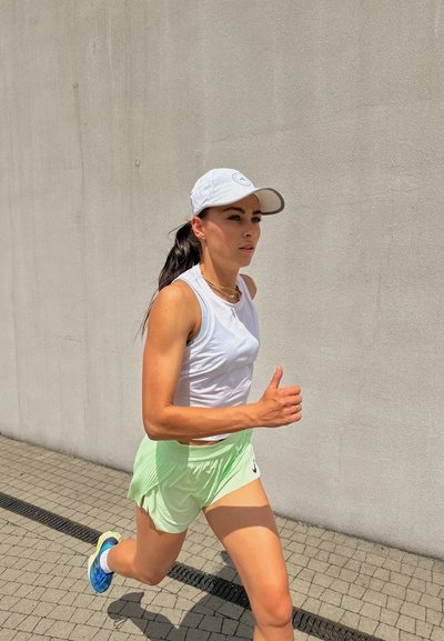 Corredor con una camiseta sin mangas blanca y pantalones cortos verde claro, lleva una gorra azul claro y zapatillas de correr azules, sobre un pavimento texturizado.