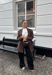 Brun oversized blazer, hvit grafisk t-skjorte, svarte vide bukser med hvite striper, brune sneakers, liten mørk veske, holder en drink.