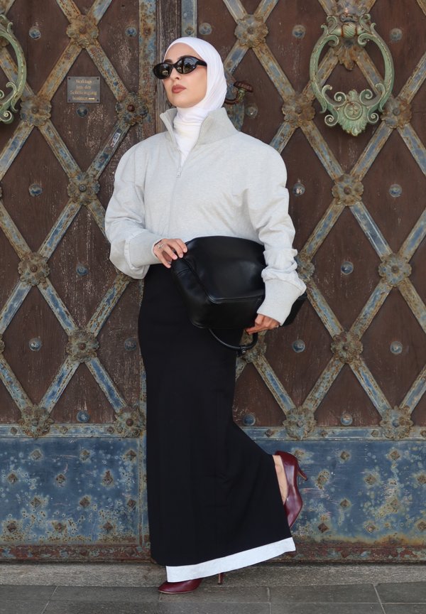 Femme portant un hijab blanc, une veste gris zippée, une jupe noire, des talons bordeaux, un sac noir et des lunettes de soleil, debout contre une porte métallique ornée.