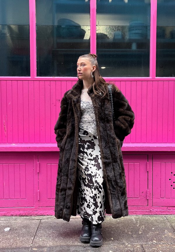 Manteau en fausse fourrure marron, haut en dentelle blanche, jupe à motif vache noir et blanc, bottes noires épaisses, posé contre un fond en bois rose.