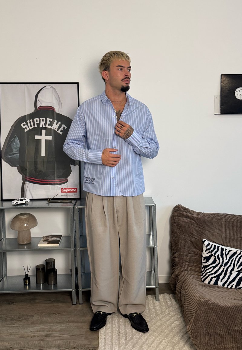 Homme avec des tatouages et les cheveux décolorés portant une chemise rayée et un pantalon beige, debout dans le salon à côté d'une étagère avec des décorations et une photo encadrée d'une veste.
