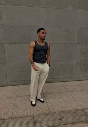 Top gris sin mangas, pantalones de pierna ancha crema y mocasines en blanco y negro. De pie contra una pared gris texturizada con un bolso de hombro.