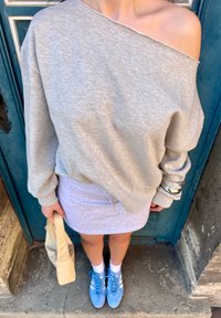 Sweatshirt cinza descaído nos ombros, saia justa cinza claro, sapatos azuis com detalhes brancos, segurando uma bolsa bege, em pé sobre uma superfície de pedra.