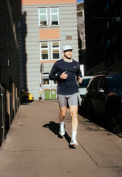 Corredor con una camiseta de manga larga azul oscuro y pantalones cortos grises, llevando una gorra clara y zapatillas blancas, trotando por la acera de una ciudad.