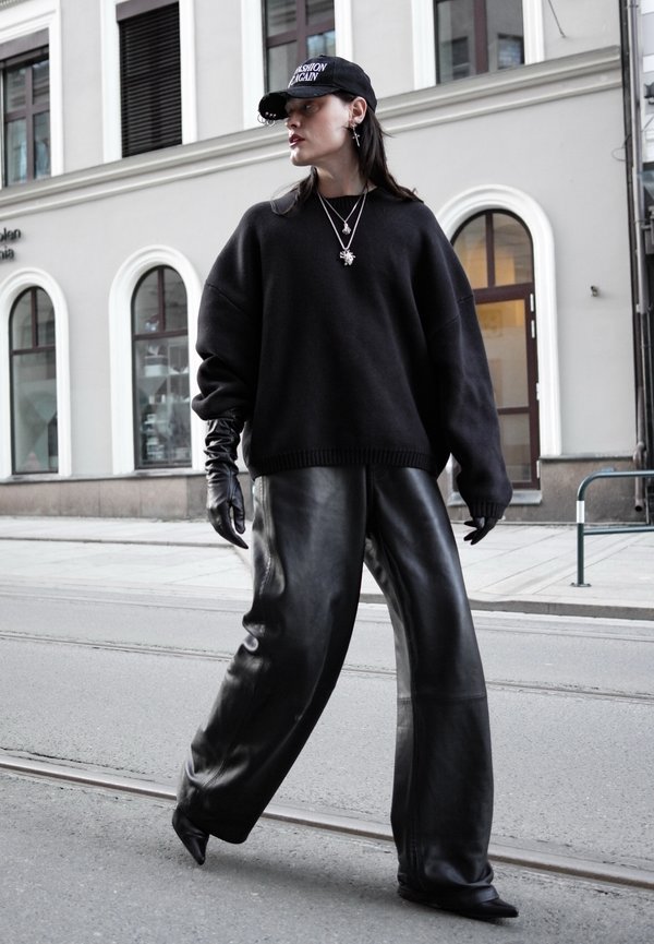 Donna che indossa un maglione nero oversize, pantaloni in pelle neri a gamba larga, guanti neri, cappellino nero con scritta, che cammina in una strada cittadina con edifici sullo sfondo.