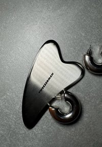 Outil de gua sha en acier inoxydable en forme de cœur courbé, avec une finition brossée et gravé "TweezerMan" ; deux crochets en métal inclus.
