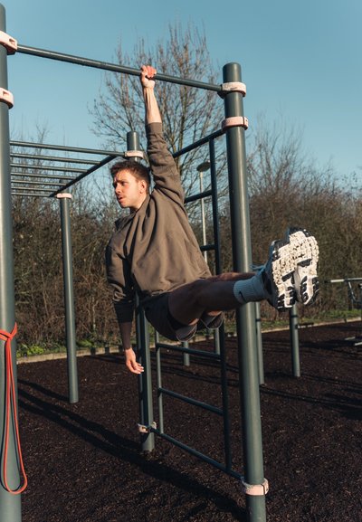 Un aparato de fitness con barras de metal y empuñaduras de goma, una persona colgando de una barra con un sudadera gris y pantalones cortos, demostrando fuerza.