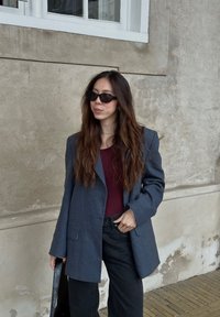 Blazer oversized cinza com mangas longas, usado sobre um top bordô, combinado com jeans largos pretos e óculos de sol pretos. Fundo de parede texturizada.