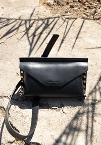 Schwarze Leder-Clutch mit einem gefalteten Klappen-Design, goldenen Nietenakzenten auf einer Seite und einem abnehmbaren Trageriemen für Crossbody- oder Handtrageoption.