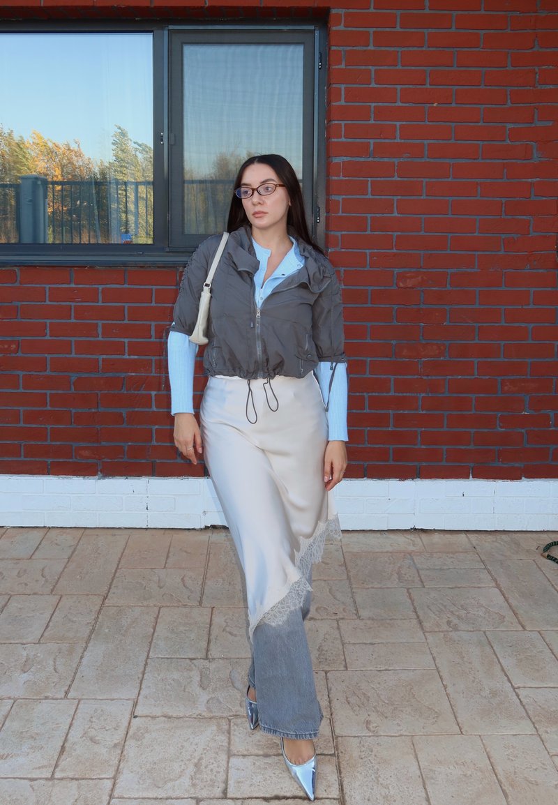 Prillidega naine, kes kannab halli jopet helehalli särke, valget asümmeetrilist seelikut, mille all on hallid teksad, ja hõbedasi terava ninaga kontsakingi, seisab punastest tellistest müüri kõrval.