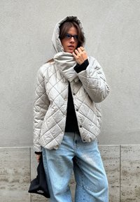 Femme portant une veste matelassée gris clair avec capuche relevée, jean bleu, lunettes de soleil, tenant un sac noir, debout contre un mur gris texturé.