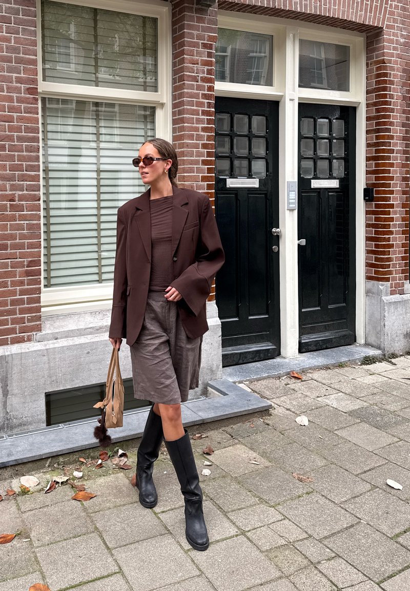 Bruine oversized blazer met een textuur bovenkant, grijze shorts, zwarte kniehoge laarzen en een beige tas met pompon-detail; stedelijke achtergrond.