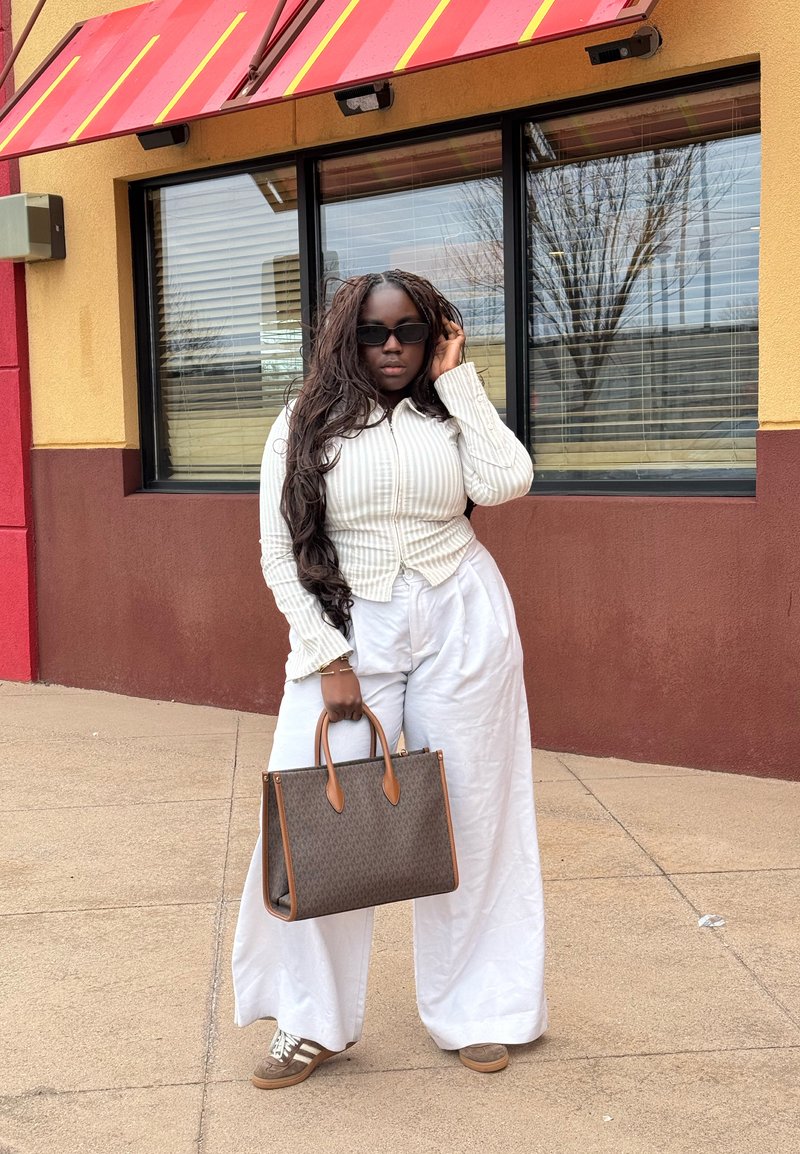Femme avec de longues tresses portant des lunettes de soleil, une veste blanche rayée, un pantalon blanc ample, des baskets marron, tenant un sac à main marron, debout devant un bâtiment.