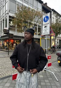 Fekete, rövid fazonú, zipzáros pulóver oldalsó zsebekkel, egy textúrált fekete felső felett viselve, világosszürke nadrággal párosítva. Külső városi környezet.