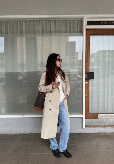 Trinchera beige de doble botonadura, suéter blanco, jeans azul claro, zapatos negros, llevando una bolsa oscura con acentos marrones, de pie junto a una ventana de vidrio.