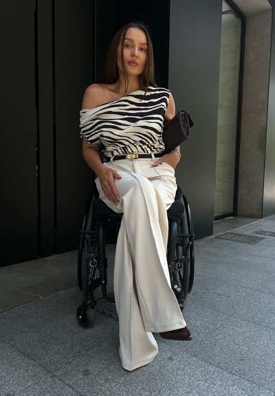 Top off-the-shoulder de rayas en blanco y negro, pantalones anchos beige, clutch negro, sentada en una silla de ruedas sobre pavimento gris.