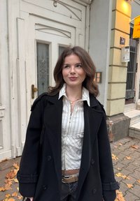 Fekete kétsoros kabát fehér csíkos inggel, látható szív alakú nyaklánccal és világos színű övvel, semleges háttér előtt.