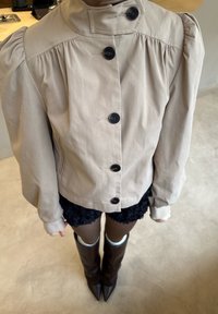 Veste beige avec épaules bouffantes, six boutons noirs et poches lat érales. Portée avec une jupe noire texturée et des bottes marron hautes jusqu'aux genoux à bout pointu.