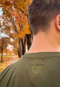 Ελιά πρασίνου μπλούζα με στρογγυλή λαιμόκοψη, με ραμμένο στρογγυλό λογότυπο σε πιο ανοιχτό πράσινο στην άνω πλάτη. Μαλακή υφή από βαμβάκι.