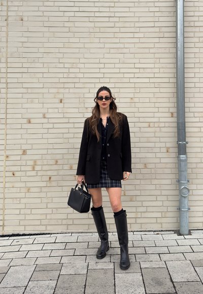 Mujer con blazer negro, falda a cuadros, botas hasta la rodilla, gafas de sol, sosteniendo un bolso negro, de pie frente a una pared de ladrillos beige con una tubería de metal.
