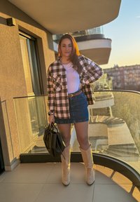 Černobílá kostkovaná flanelová košile, bílé tričko, džínová sukně a béžové kozačky po kolena. Drží černou kabelku. Prostředí venkovního balkonu.