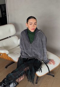 Jeune femme aux cheveux lisses assise sur une chaise moderne blanche, portant un pull gris à fermeture éclair, un col roulé vert, une jupe à carreaux et des bottes noires.