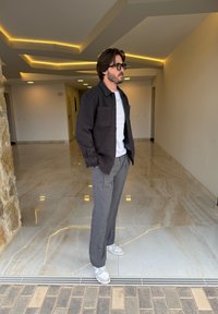 Chaqueta abotonada de lana gris oscuro sobre una camiseta blanca; pantalones a rayas grises; zapatillas blancas; de pie en un espacio interior bien iluminado.