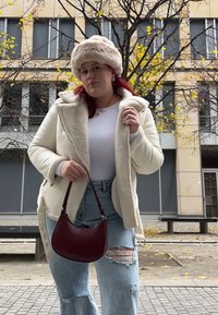 Κρέμα-χρώματος τζάκετ από γούνινο υλικό με φαρδύ γιακά, λευκό μπλουζάκι, φθαρμένες μπλε τζιν, και μπορντό τσάντα. Χαρακτηριστικά περιλαμβάνουν κουμπιά στο τζάκετ.