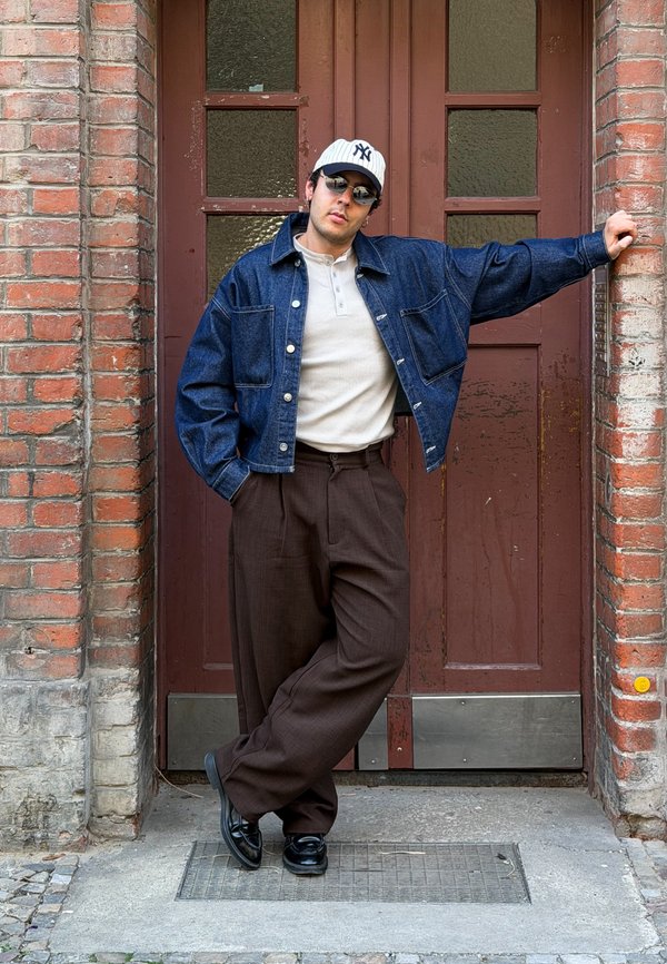 Mann mit Jeansjacke, weißem Henley-Shirt, brauner Hose, Sonnenbrille und weißer Baseballkappe lehnt mit überschlagenen Beinen an einem gemauerten Türrahmen.